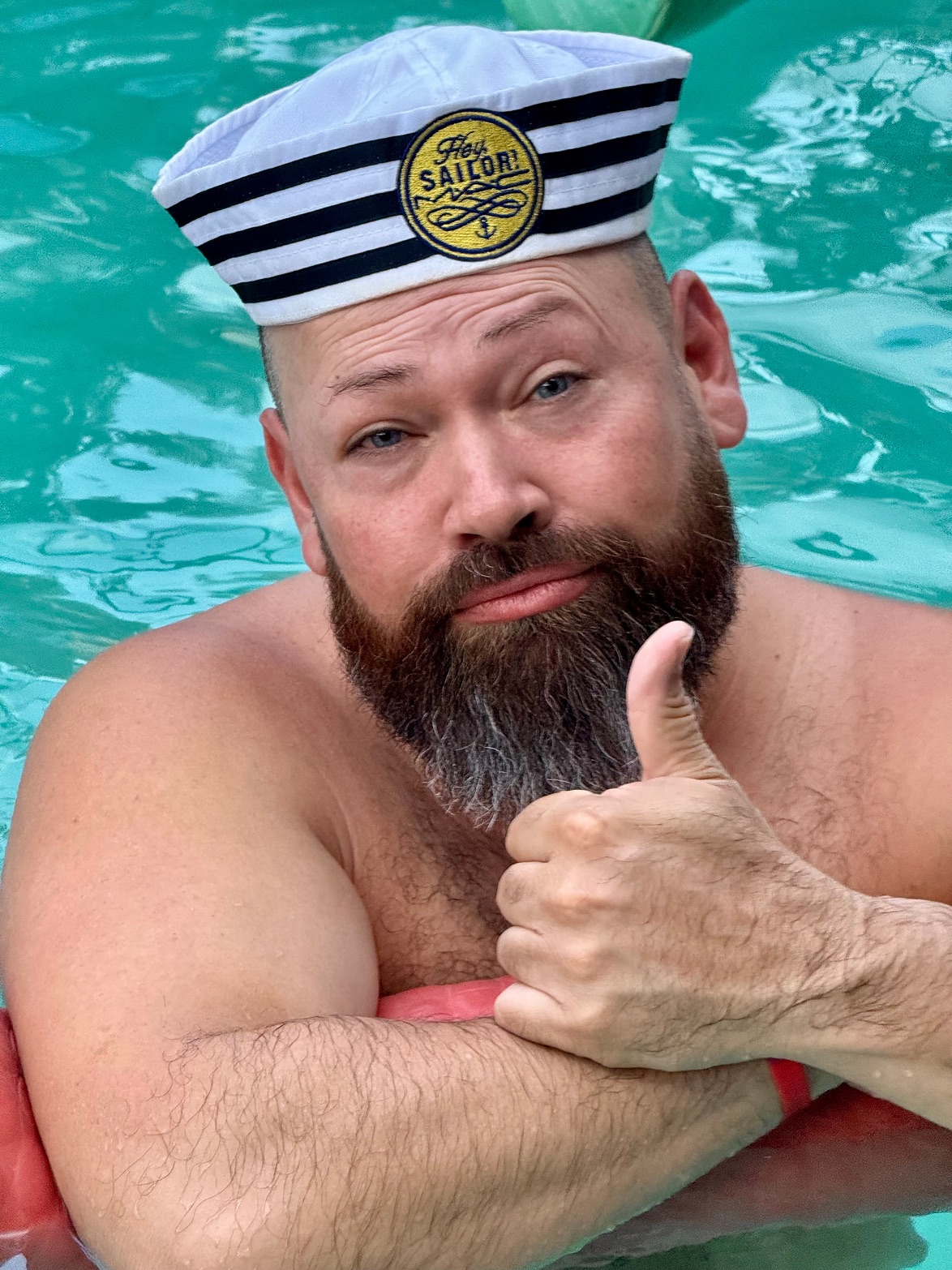Bearded man in a sailor hat giving a thumbs-up in a pool