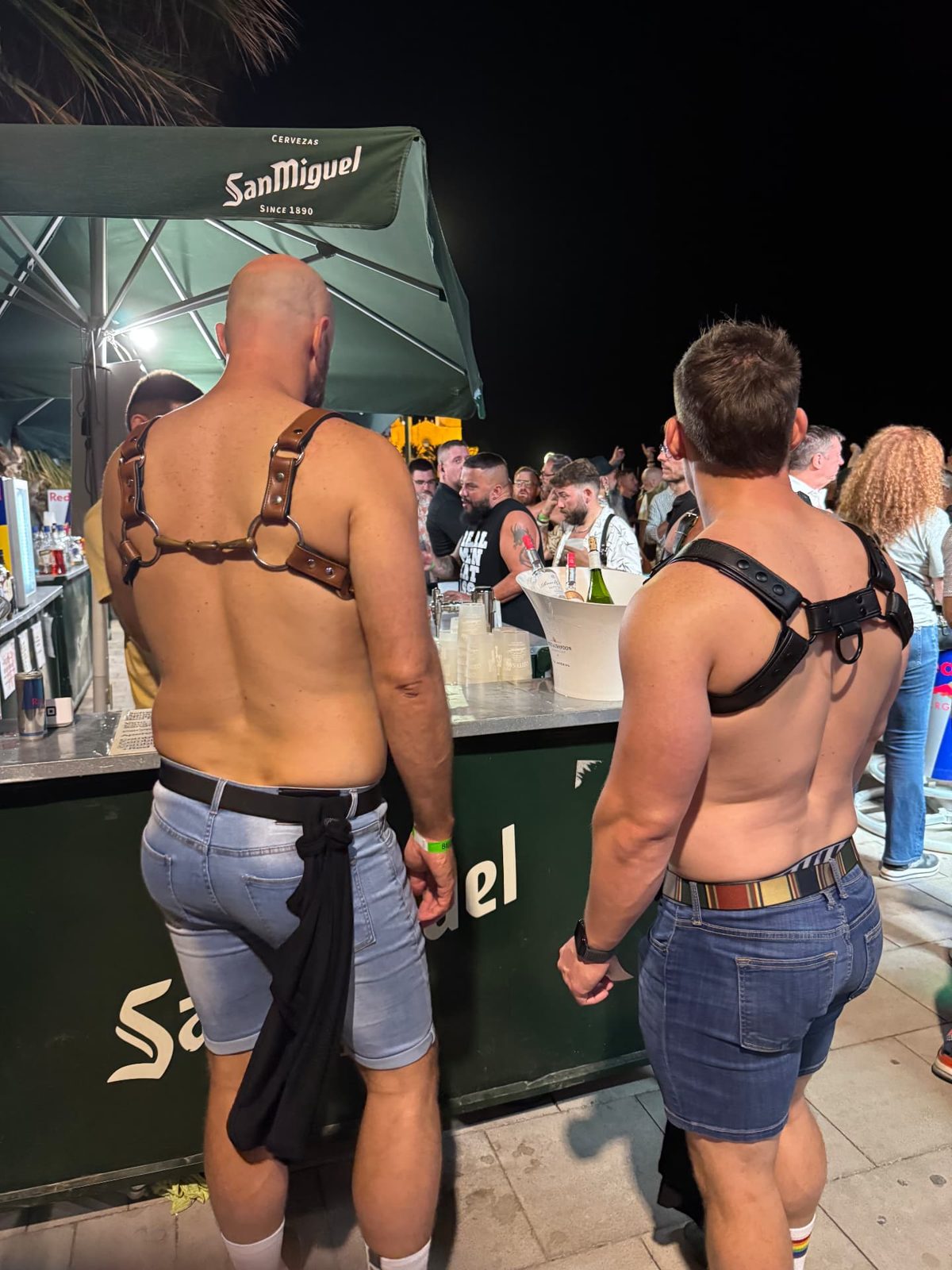 Two bears in leather harnesses ordering at an outdoor bar in Sitges